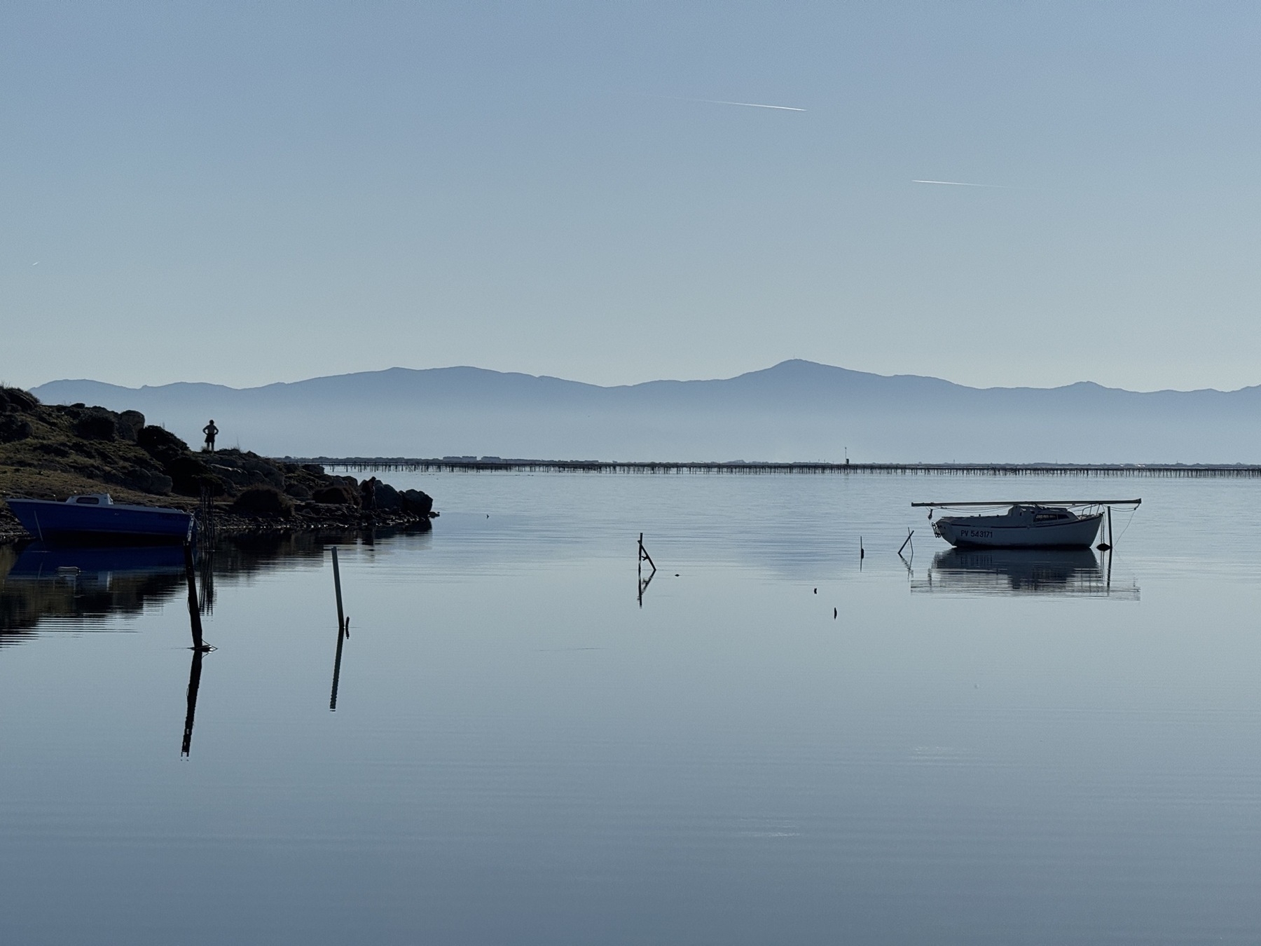 A serene lakeside scene features boats on calm water with distant mountains in the background.