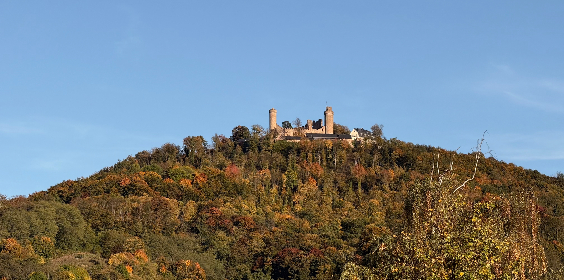 Auerbach Castle, with multiple towers, stands atop a tree-covered hill with autumn foliage under a clear blue sky.