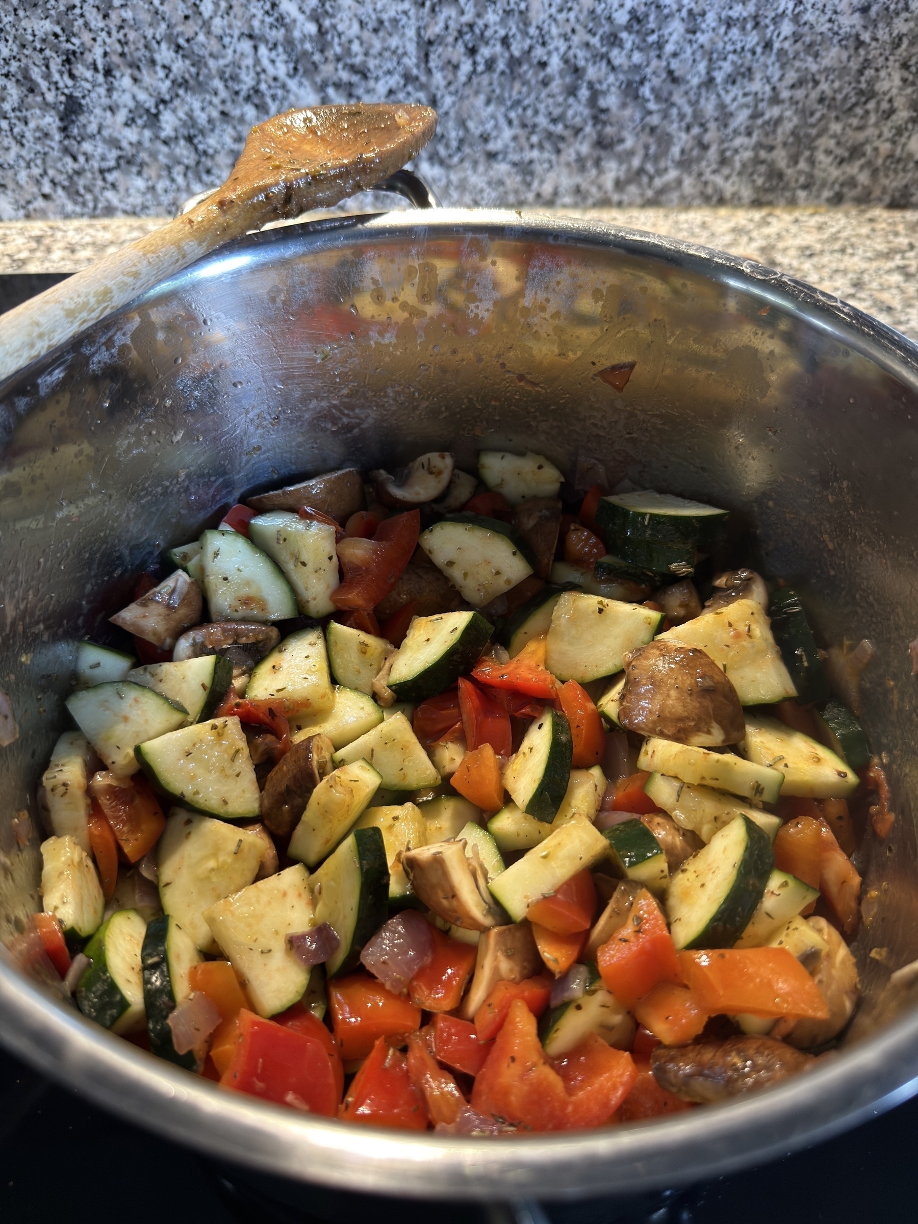 A pot filled with a colorful mix of chopped vegetables including zucchini, mushrooms, and red bell peppers is being cooked, with a wooden spoon resting on the edge.