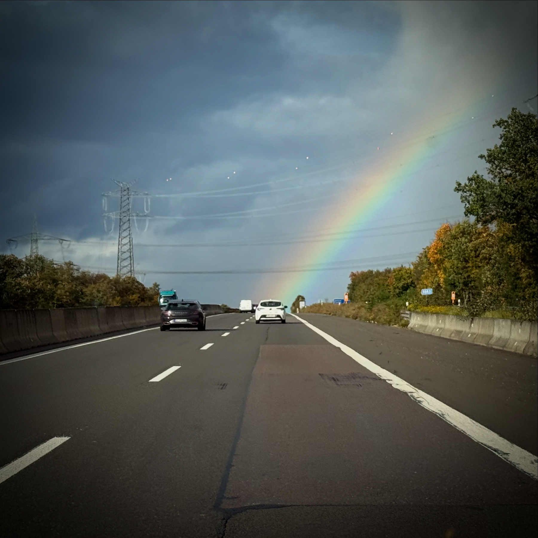A rainbow arcs over a highway with several cars driving under a partly cloudy sky.