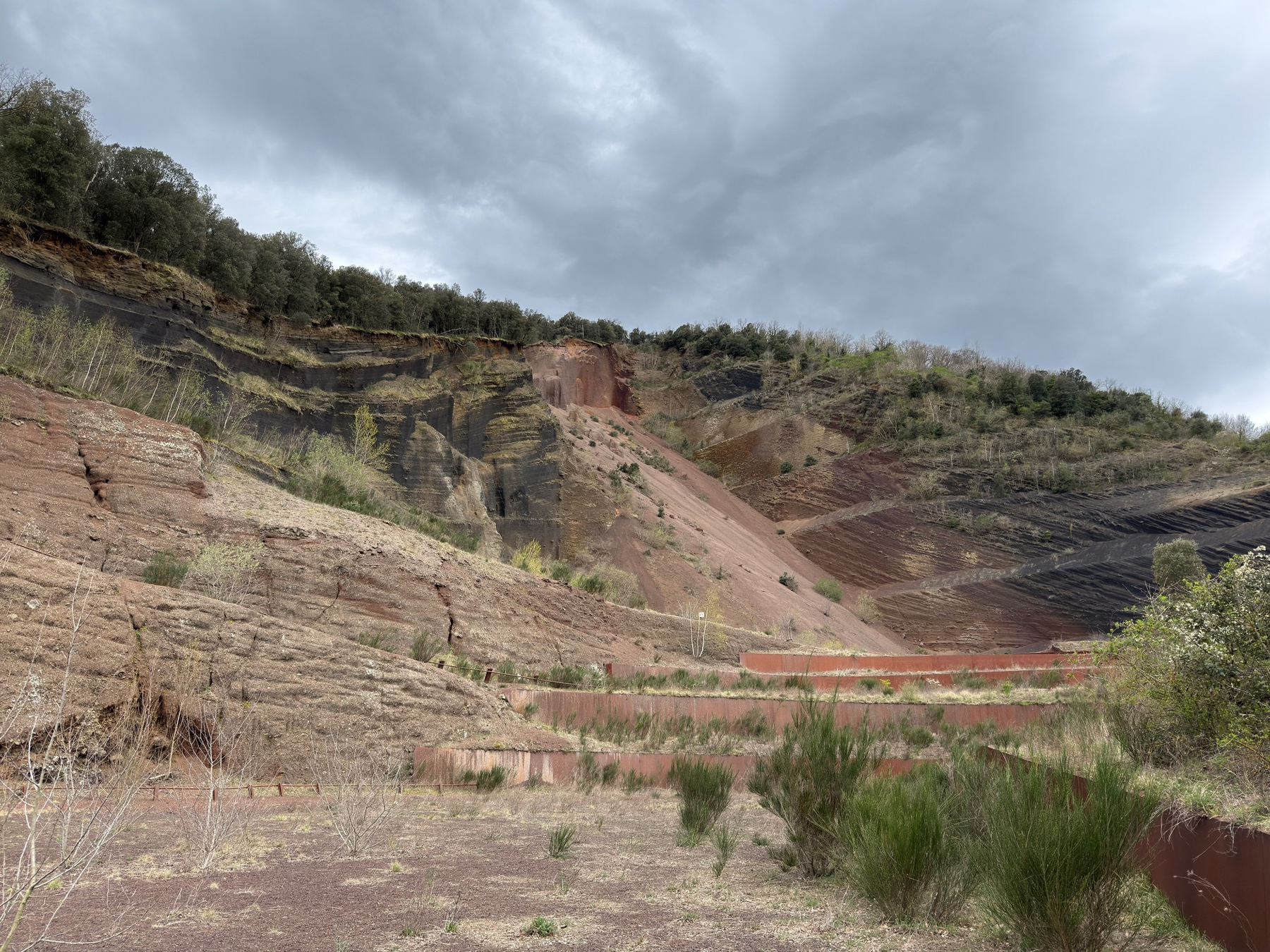 A dramatic, stratified quarry landscape is dominated by red and brown earth tones under a cloudy sky. This is the excavated side of the Volcà del Croscat