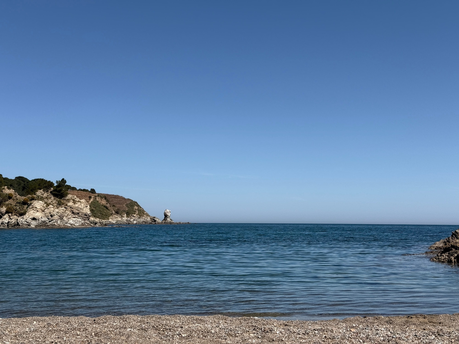 Plage des Elmes. A tranquil beach scene features a rocky coastline under a clear blue sky.