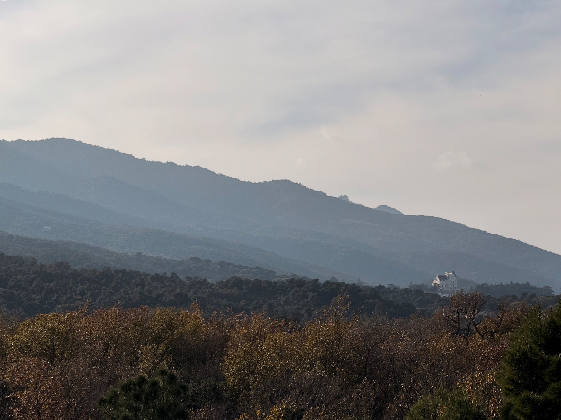 A scenic landscape features rolling hills and a distant house surrounded by autumn-colored trees under a cloudy sky. A tiny chateau is visible on the bottom right of the photo. 
