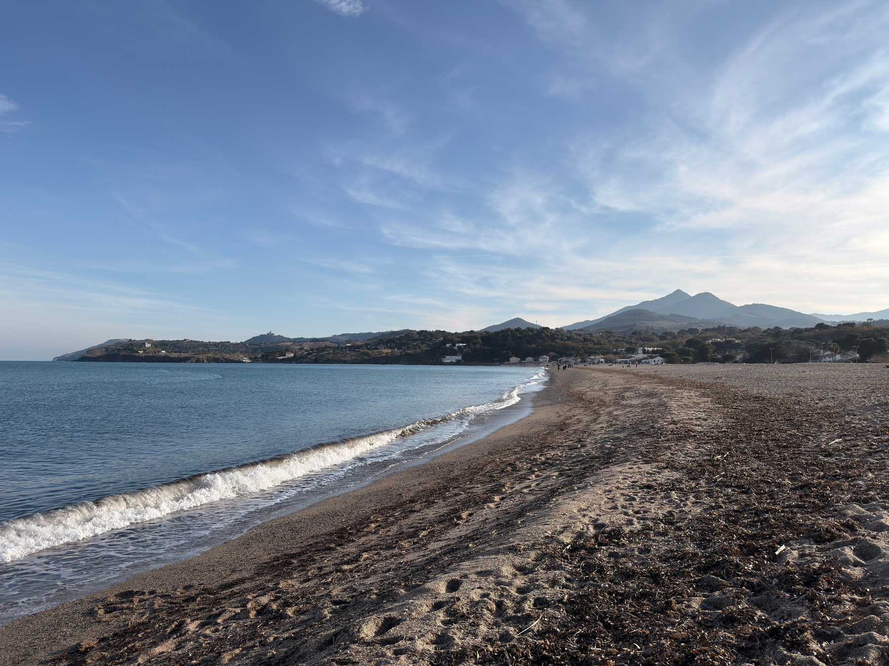 A sandy beach stretches along a calm sea with distant hills under a blue sky.