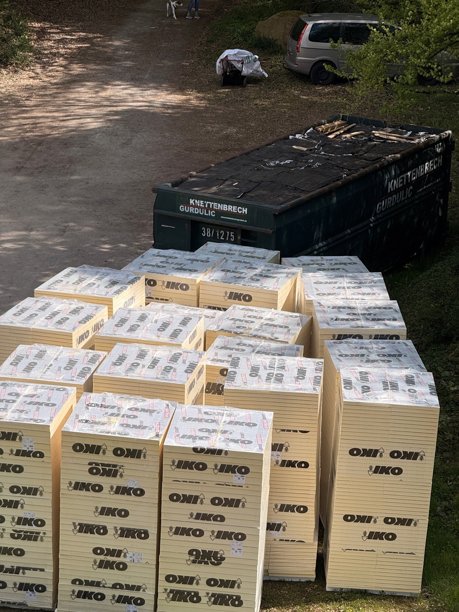 A large number of stacked boxes are arranged near a dumpster and a parked vehicle on a dirt road, with trees in the background. They look like giant blocks of Post-It notes!