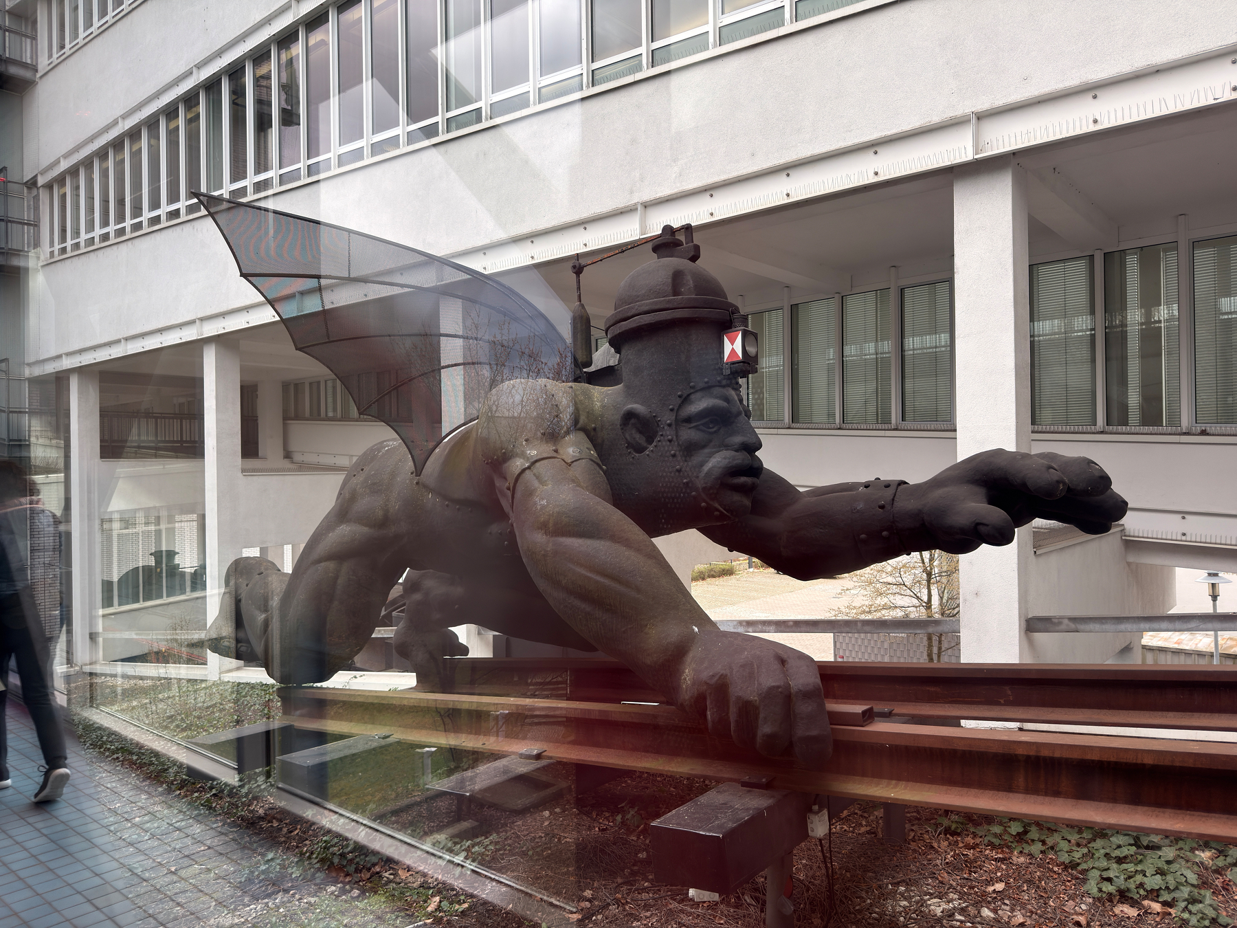 An enormous sculpture of a winged figure is displayed outside a modern building, designed to appear as if it is flying along two iron rails. &10;&10;The sculpture is called the “Maschinenmensch” (Machine Man).&10;&10;It is described as “half locomotive, half human figure, half iron devil, half iron angel” — a steam-breathing figure bound to a rail track. The museum’s own text says it represents the conflicted feelings people have had toward technology: fear on one side, hope for progress on the other.&10;&10;A Polish artist created it in 1989, based on a caricature from 1884. It is roughly 6-7 meters long and 3 meters tall. 