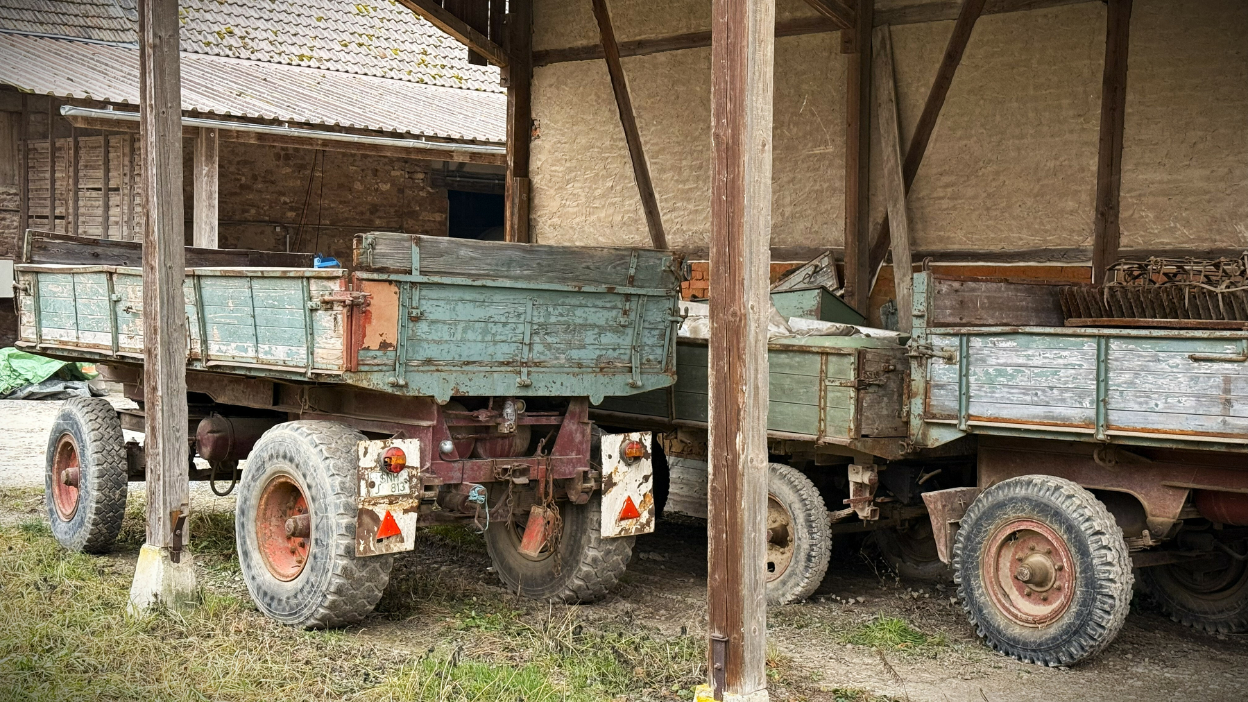 Two old, weathered trailers with large tires are parked under a wooden, open-sided shed.