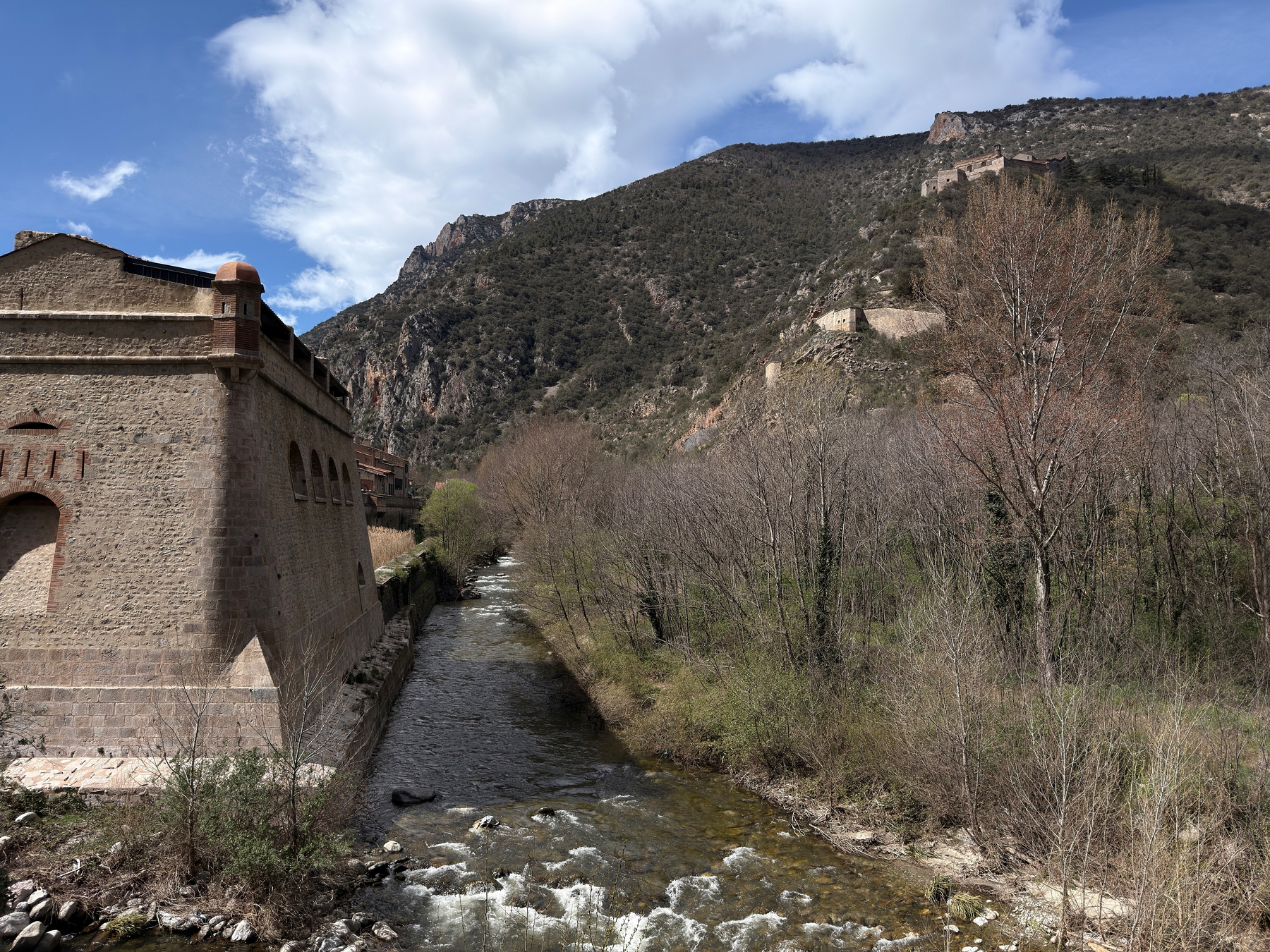 A stone fortress with rounded turrets stands beside a narrow river gorge cutting through rocky terrain. Mountains rise in the background under a partly cloudy sky. A road bridge runs parallel to the fortress wall on the right.