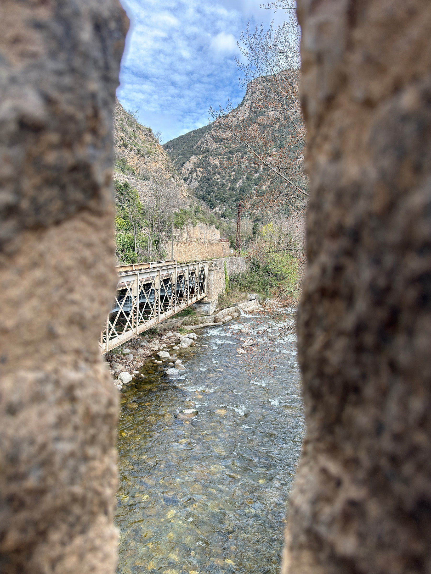 A river flows under a bridge surrounded by rocky terrain and trees, viewed through a narrow stone gap in the town walls of Villefranche-de-Conflent.