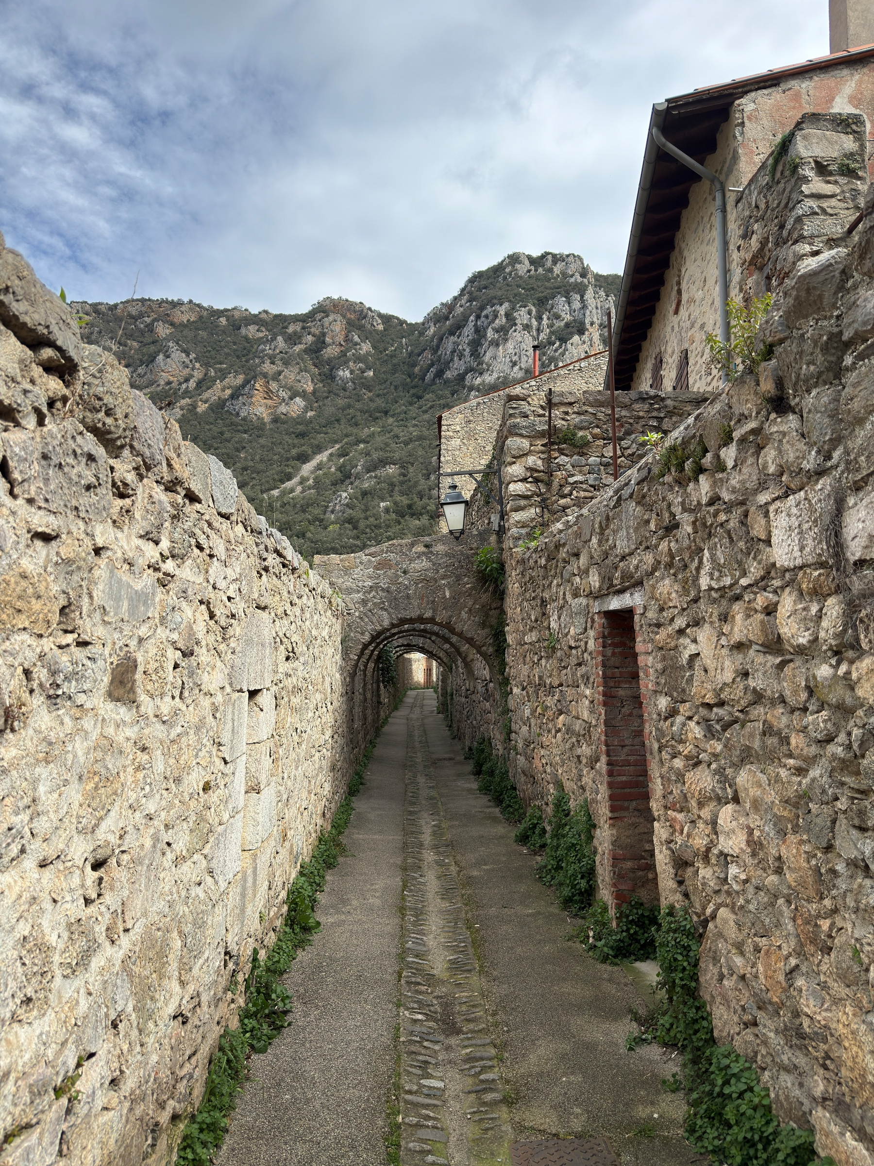 A narrow stone pathway is flanked by ancient stone walls, leading towards an archway with mountainous terrain in the background.