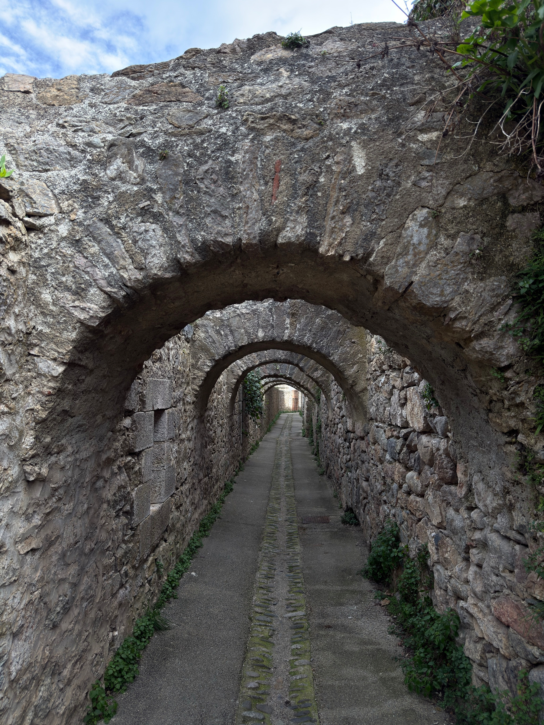 A long stone-arched passageway recedes into the distance, with each arch framing the next in a repeating pattern. The walls are rough-cut stone with patches of moss and ivy. A central drainage channel runs along the paved floor.