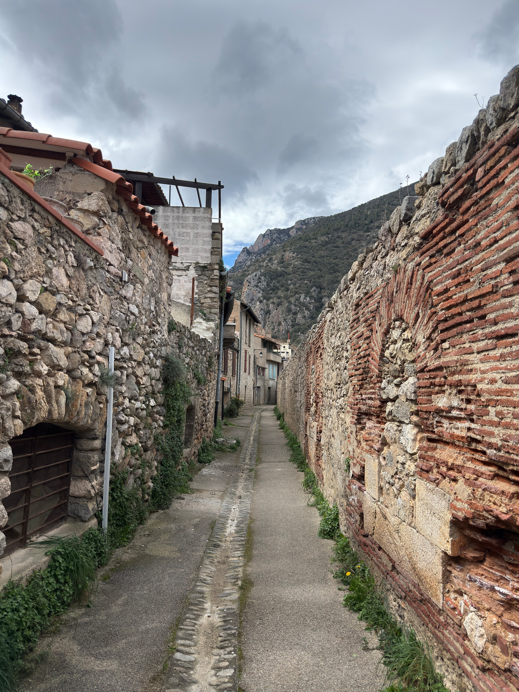 A narrow, cobblestone alleyway is flanked by rustic stone buildings and leads towards a distant mountainous landscape under a cloudy sky.