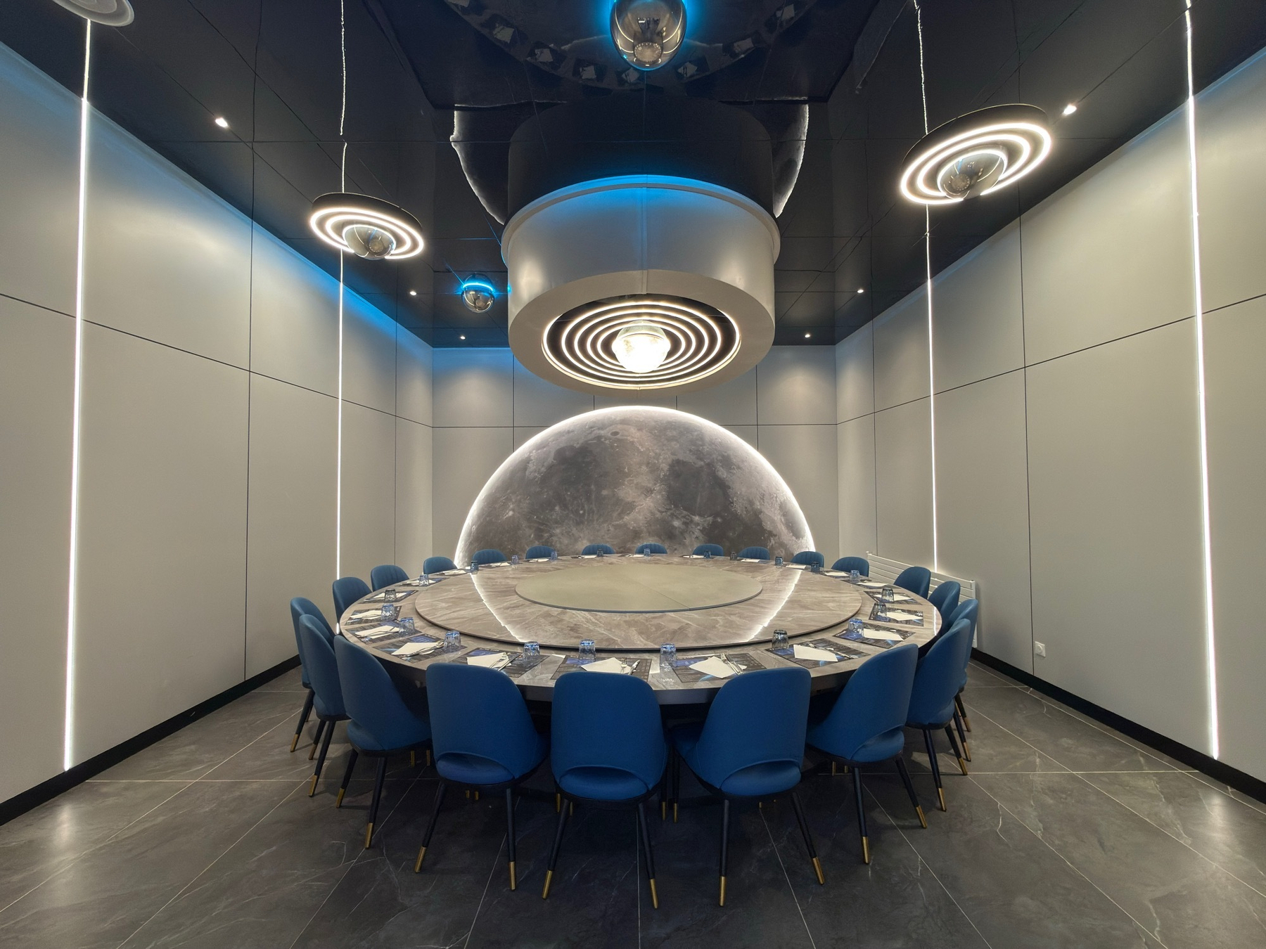 A futuristic dining room features a round table with blue chairs, illuminated by modern circular light fixtures and a moon-like backdrop.