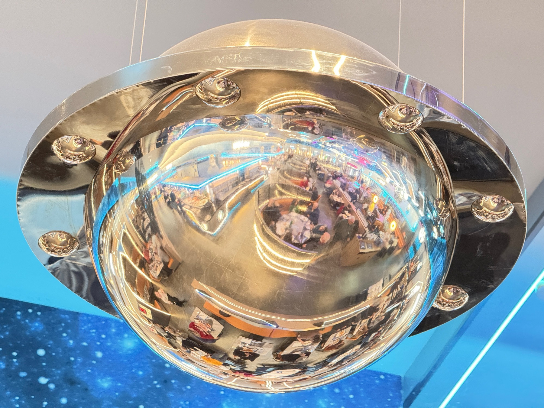 A reflective, metallic dome-shaped fixture hangs from the ceiling, displaying distorted reflections of a brightly lit interior space with people and various objects.