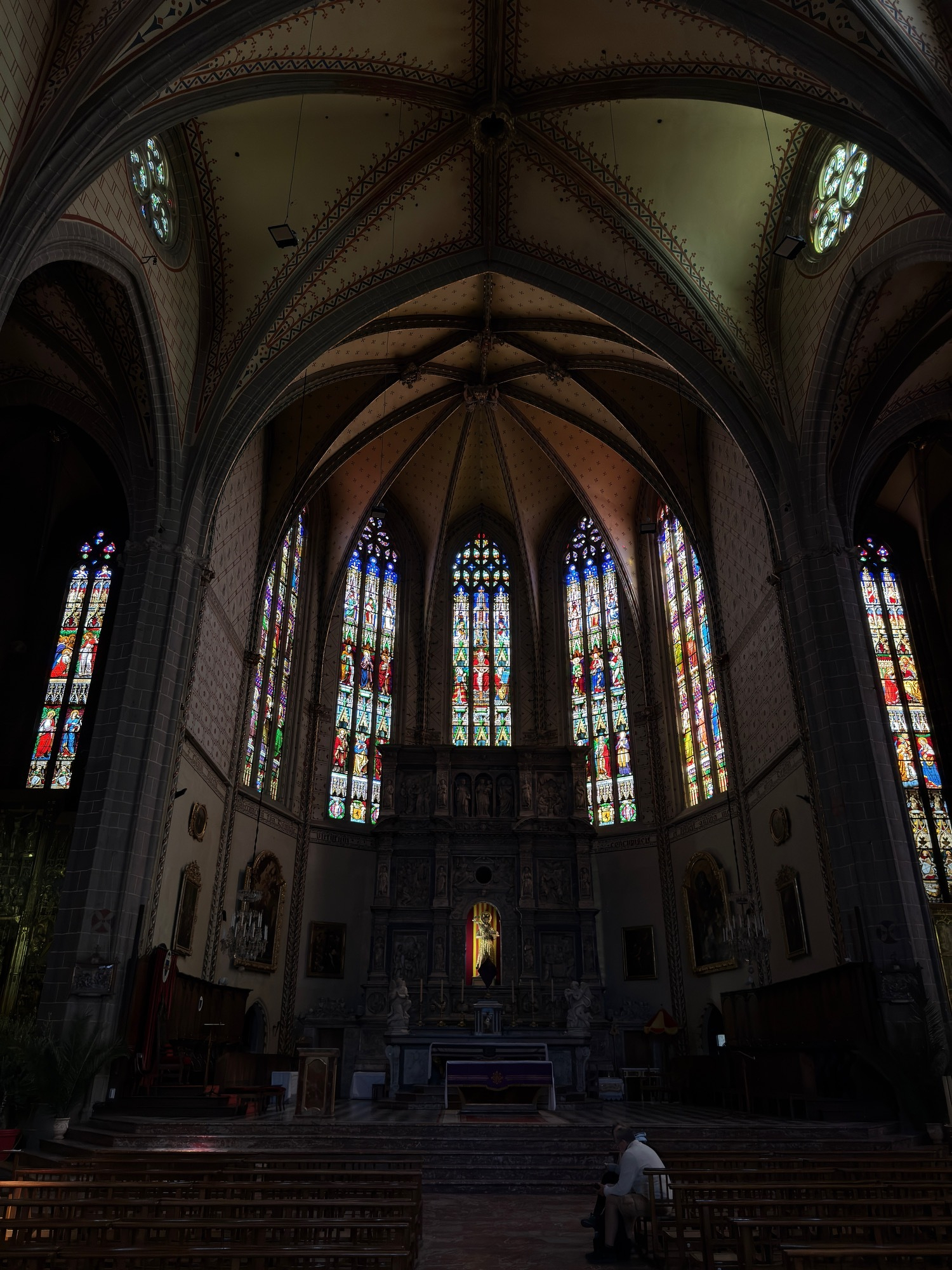 A grand cathedral interior features ornate stained-glass windows and intricate architectural details with a seating area and an altar at the center. This is the Basilique-Cathédrale Saint-Jean-Baptiste.