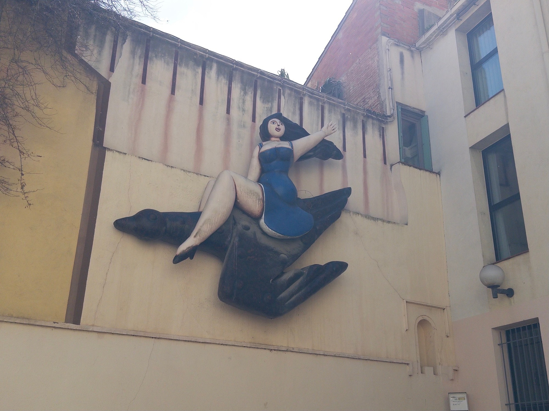 A large sculpture of a woman in a blue dress riding a bird is mounted on a building's exterior wall.